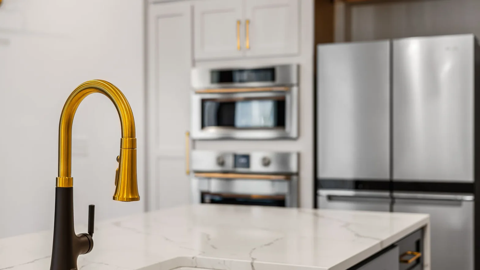 Gold curved kitchen faucet over a white marble island with stainless steel appliances in the background.