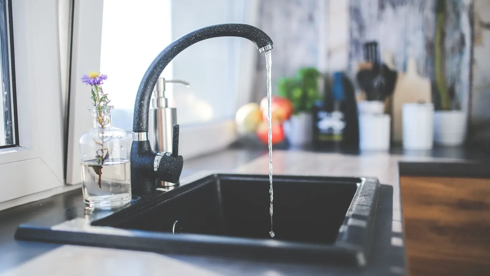 Curved black kitchen faucet pouring water into a dark sink; a glass bottle with a purple flower sits on the counter.